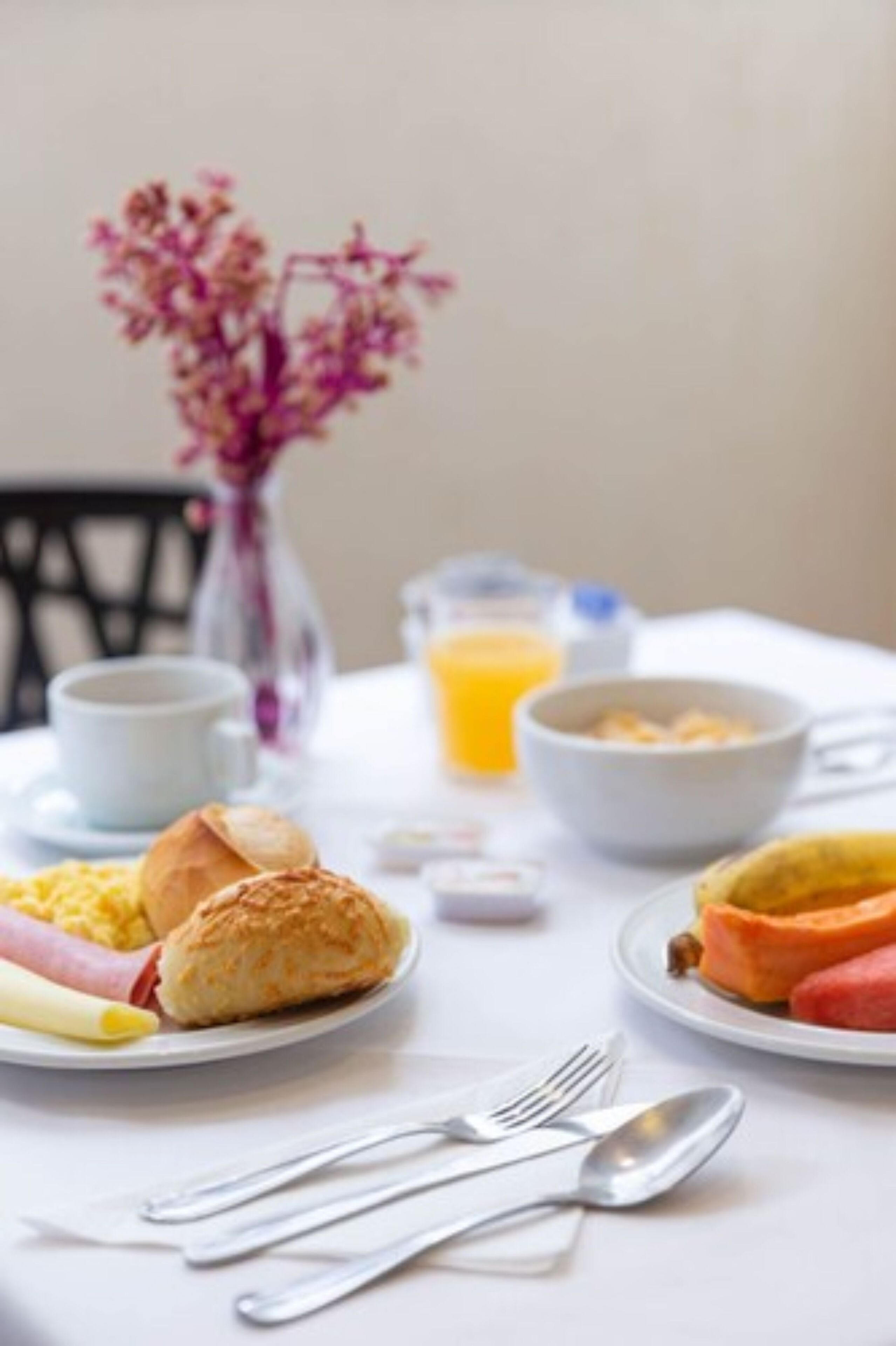 A breakfast table with croissants, a bowl of cereal, a glass of orange juice, and a vase of flowers.