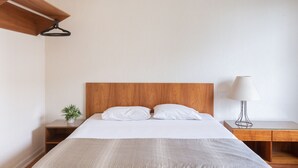 Una cama bien hecha con cabecera de madera, una mesita de noche con lámpara y una planta pequeña en el buró.