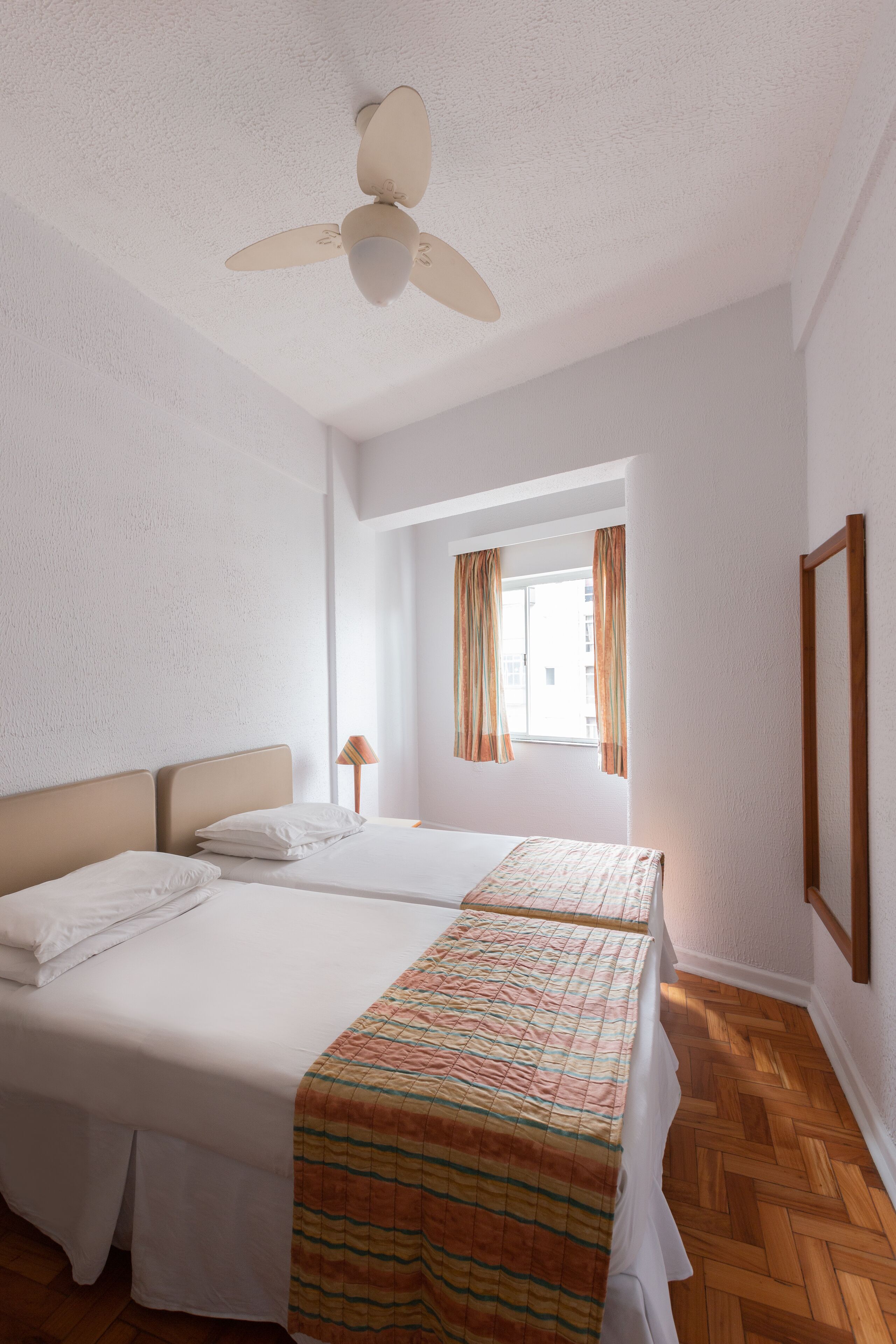A hotel room with two beds, a ceiling fan, a window with curtains, and a wooden floor.