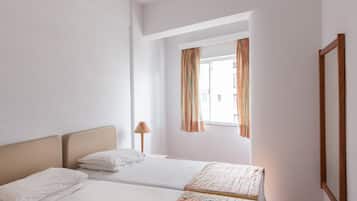 Quarto de hotel com duas camas, um ventilador de teto, uma janela com cortinas e piso de madeira.