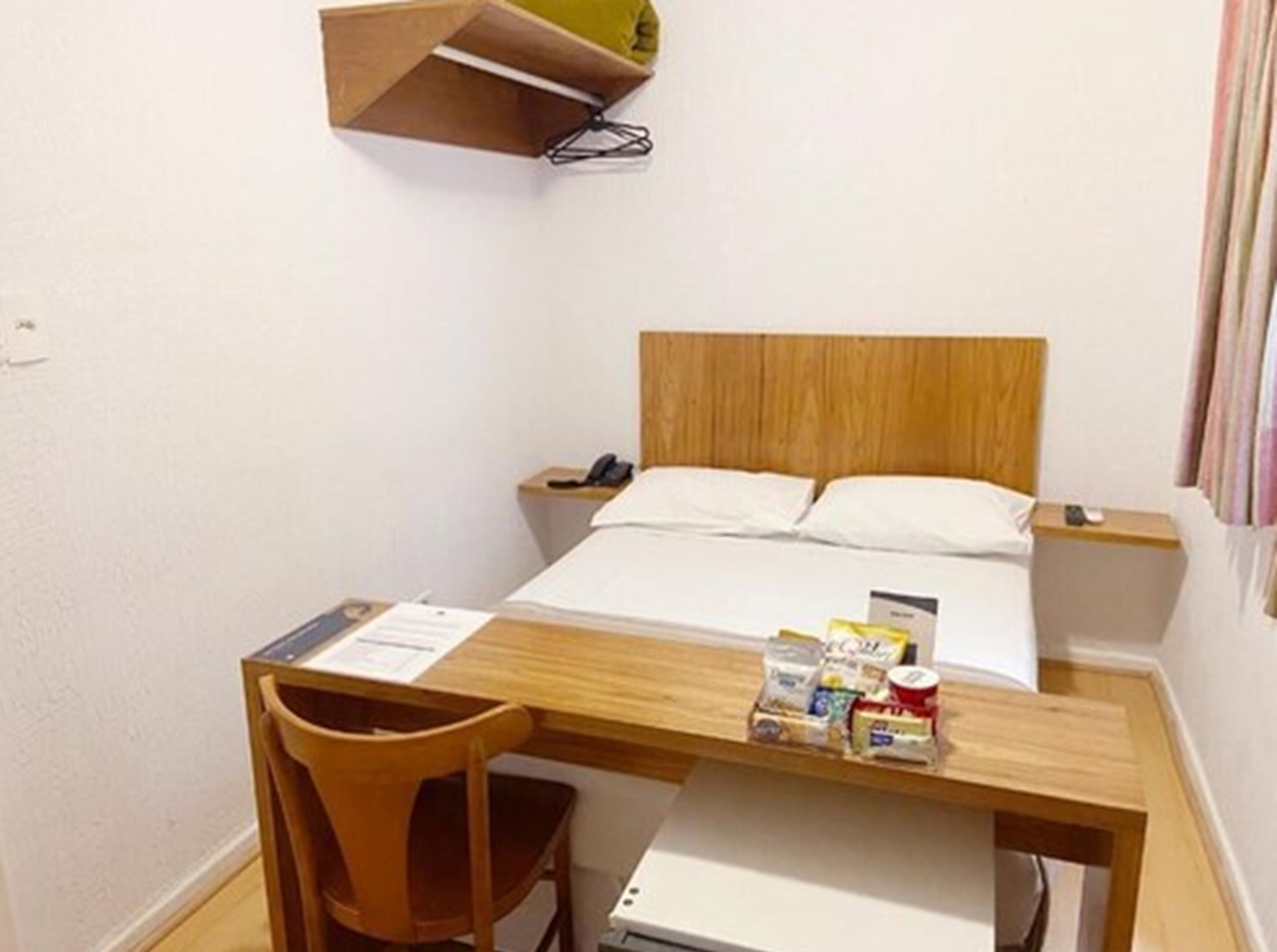 A hotel room with a bed, wooden headboard, bedside tables, a desk with a chair, and a shelf above the desk.