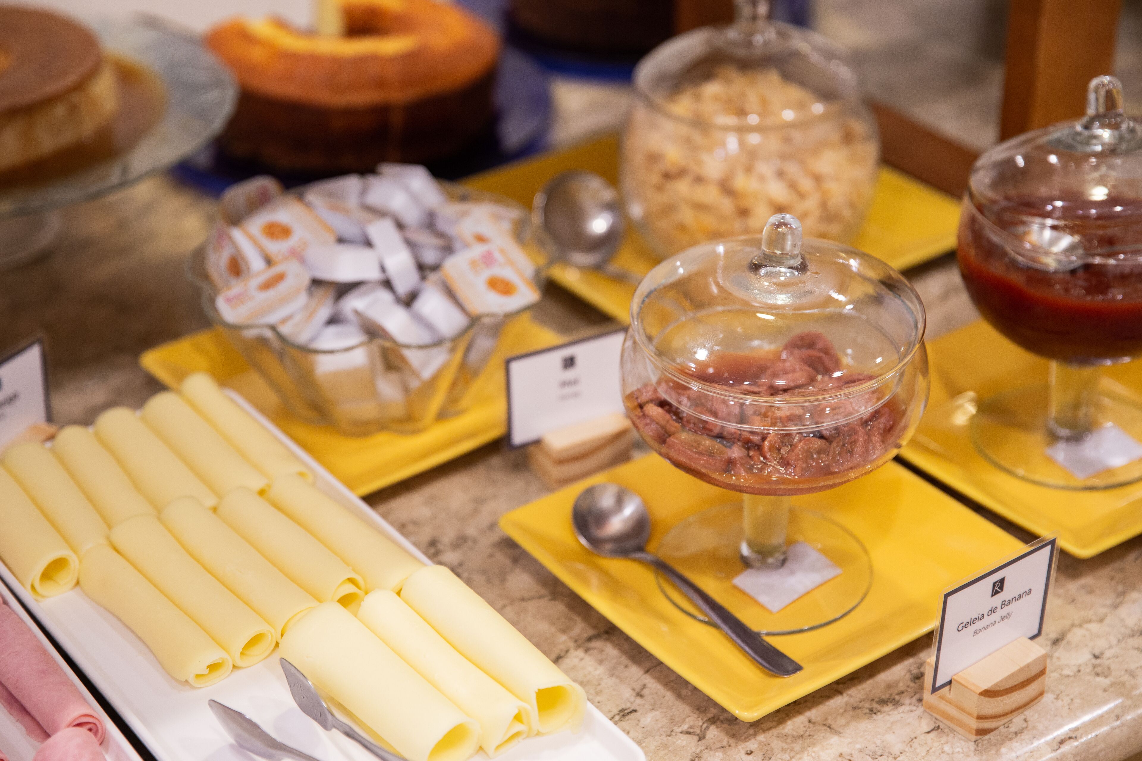 A buffet with various food items including rolled pastries, a glass container with a red sauce, and a sign indicating 'Geleia de Banana'.