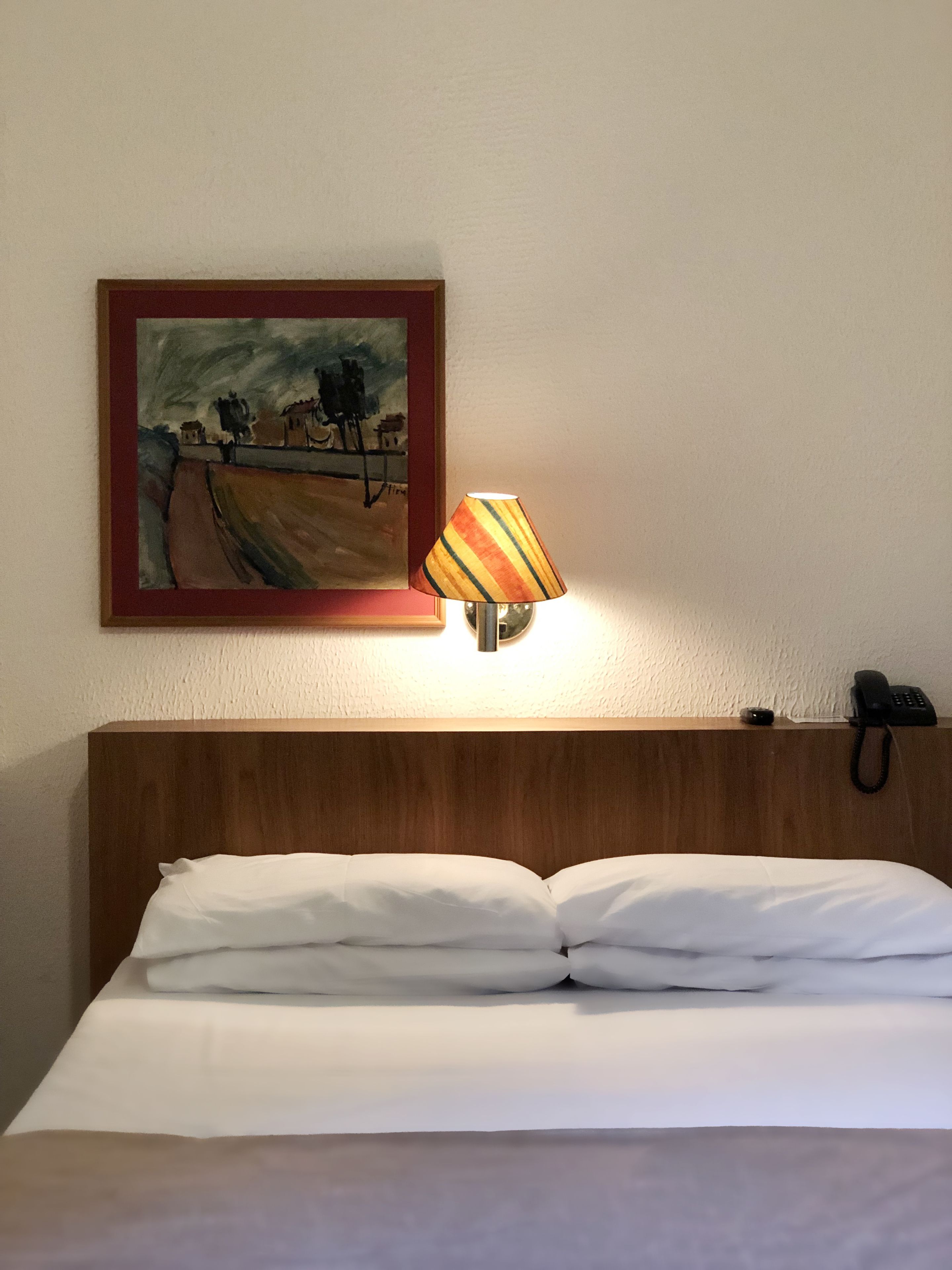A hotel room with a bed, a lamp, a painting, and a phone on the nightstand.
