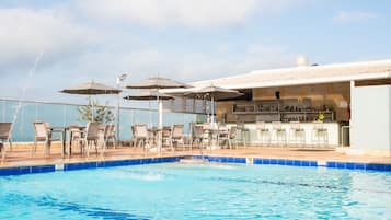 Piscina con sillones de playa y sombrillas, zona de bar con botellas y cielo despejado.