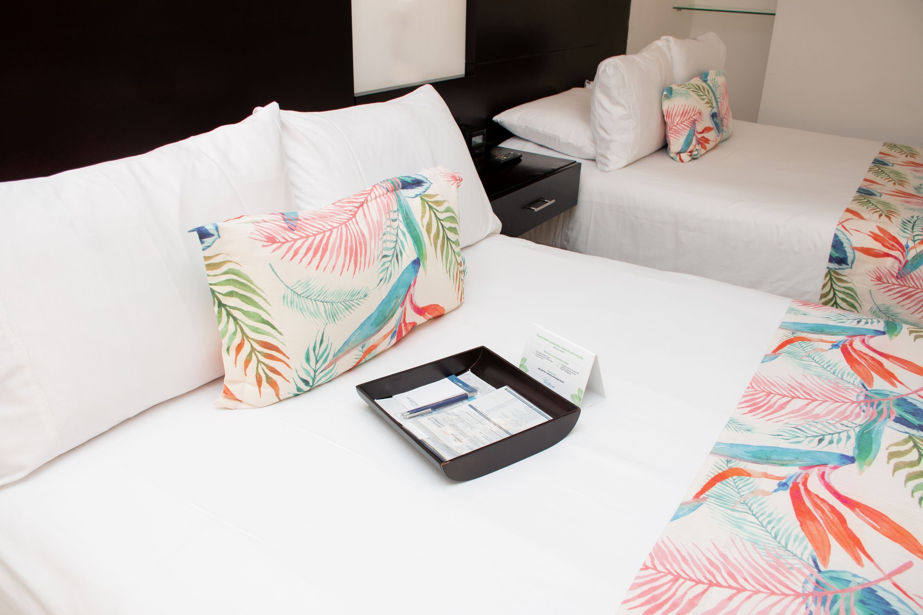 A hotel room with two beds, each with white bedding and colorful tropical leaf-patterned pillows. There is a black tray table with a pen and a business card.