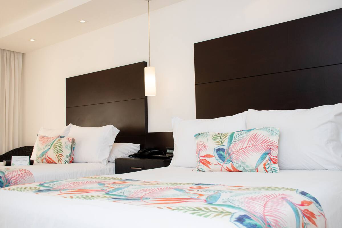 A hotel room with two beds, each with white bedding and colorful tropical leaf-patterned pillows. There is a dark headboard behind each bed, and a small bedside table with a lamp.