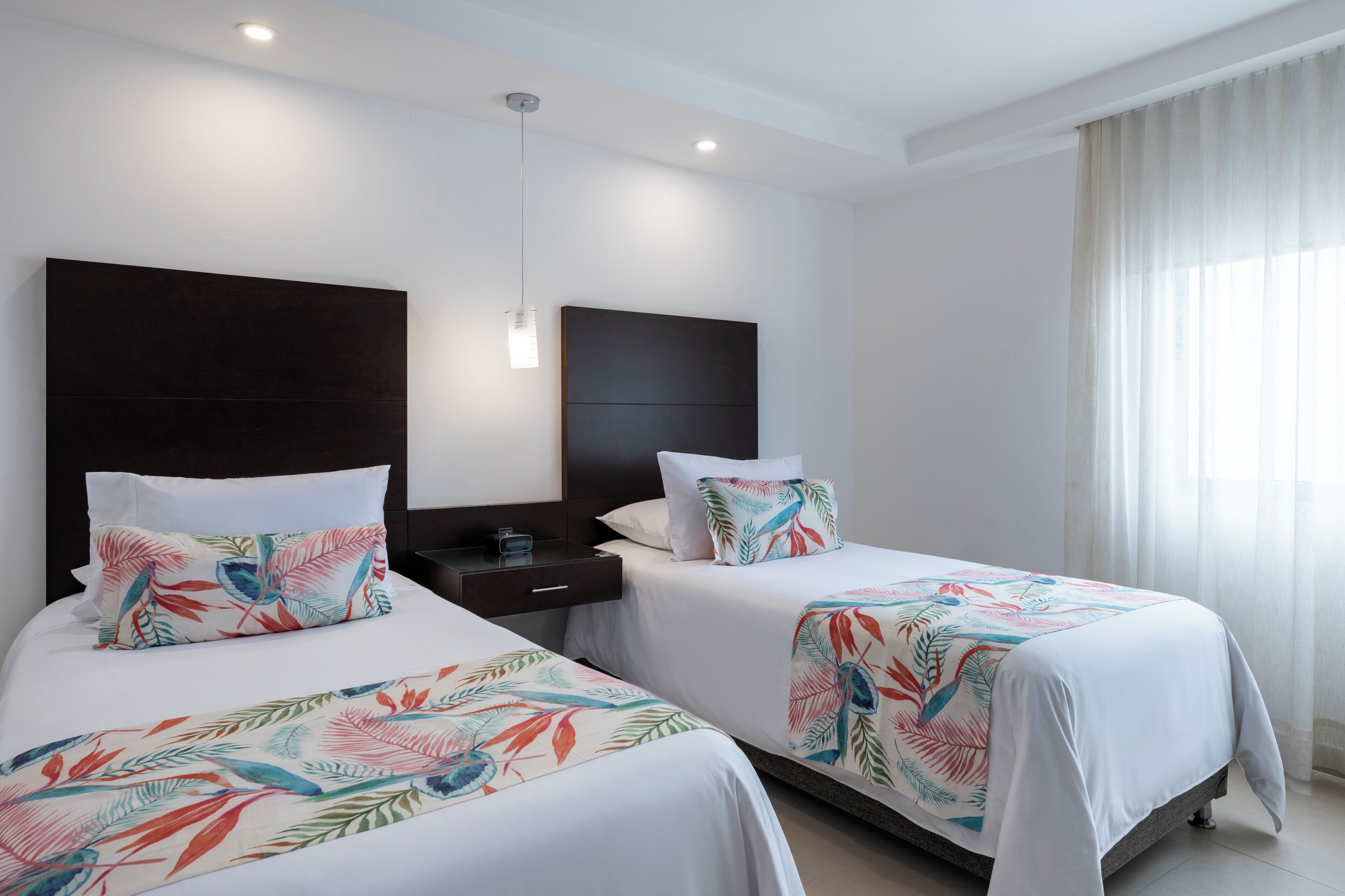 A hotel room with two beds, each with a floral bedspread, a dark headboard, a bedside table, and a nightstand.