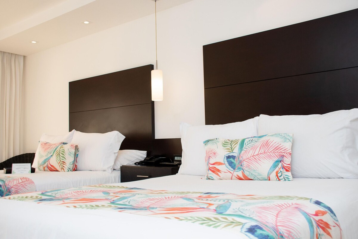 A hotel room with two beds, each with a dark headboard, white bedding, and colorful tropical leaf-patterned pillows.