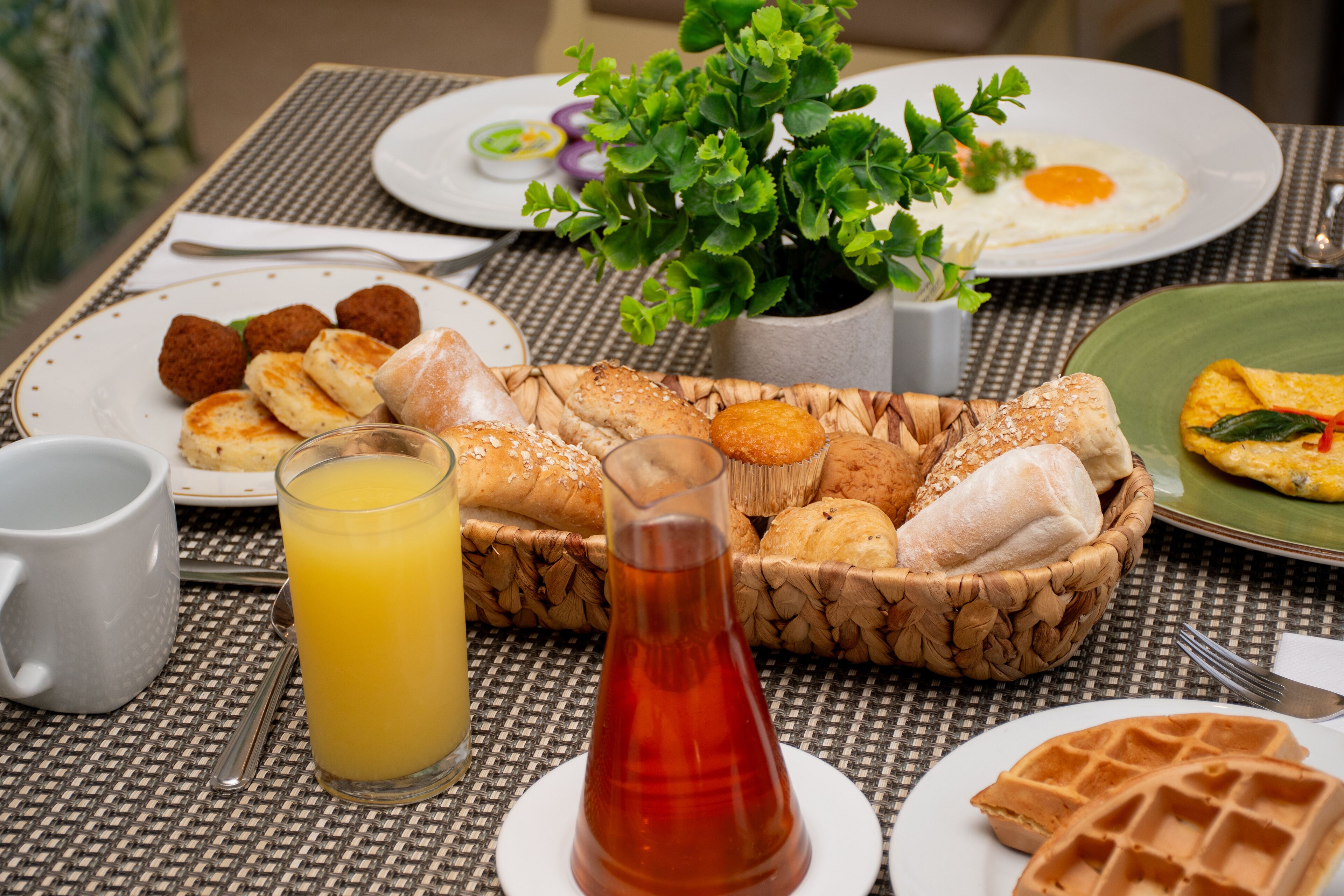 Uma mesa de café da manhã com diversos alimentos, um copo de suco de laranja e um vaso de planta.