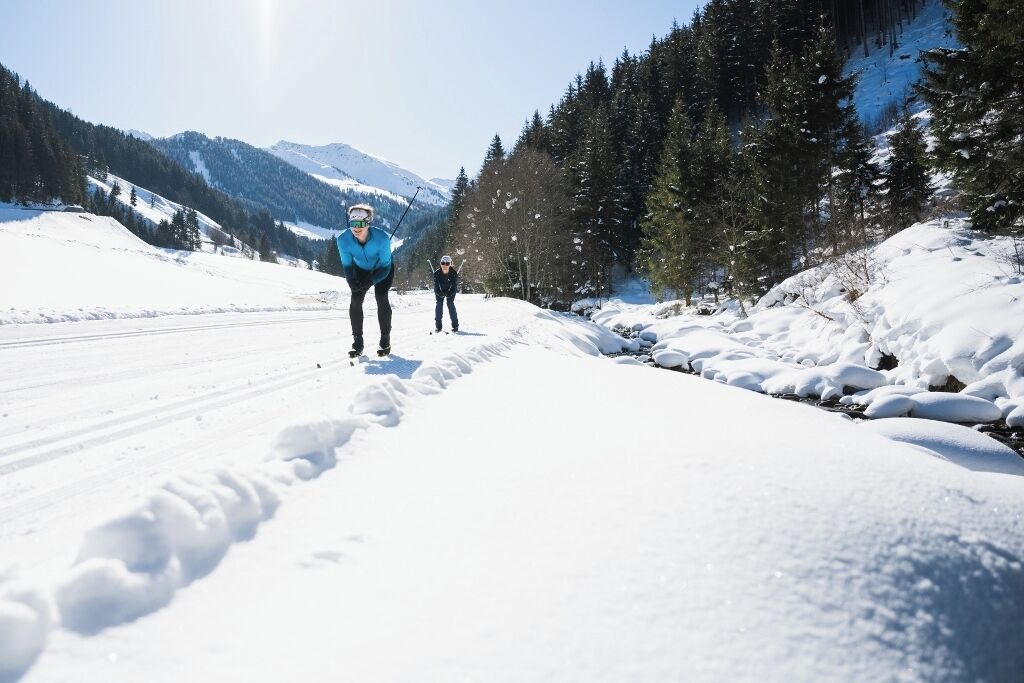 A snow-covered path with skiers, surrounded by trees and mountains.