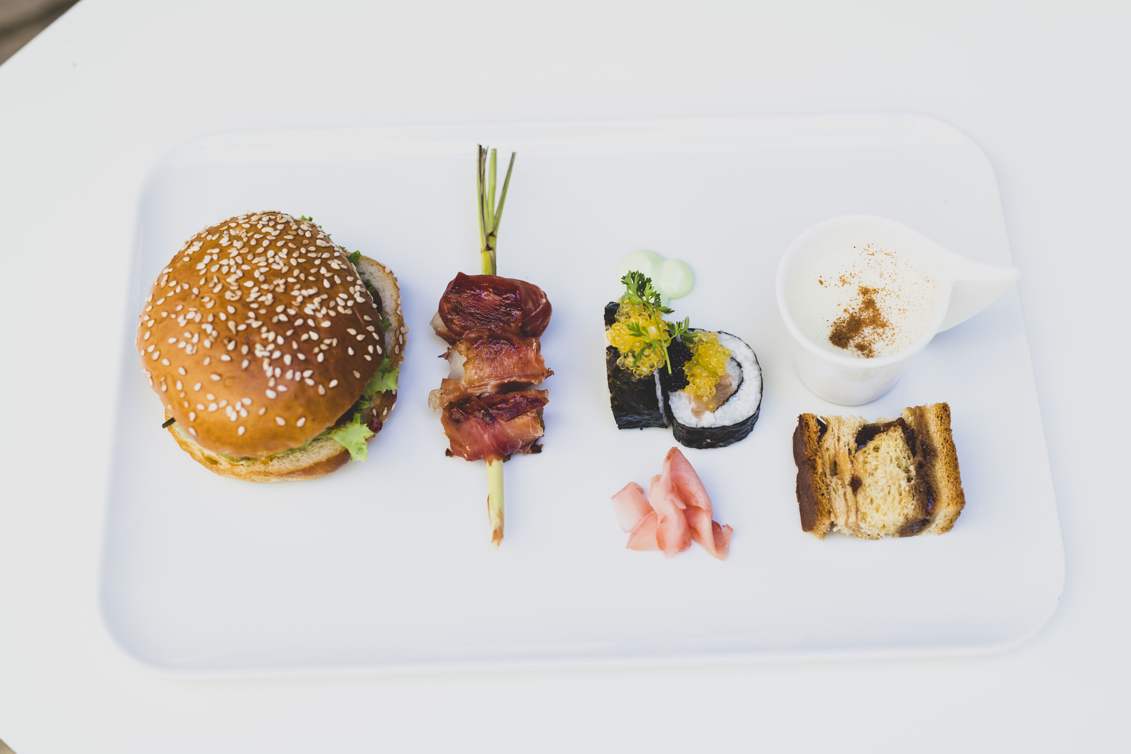 A white rectangular plate with a sesame seed bun, a bacon skewer, sushi, pickled vegetables, and a small cup of sauce.