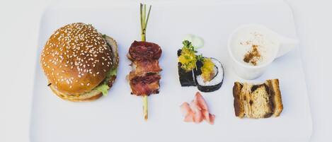 A white rectangular plate with a sesame seed bun, a bacon skewer, sushi, pickled vegetables, and a small cup of sauce.