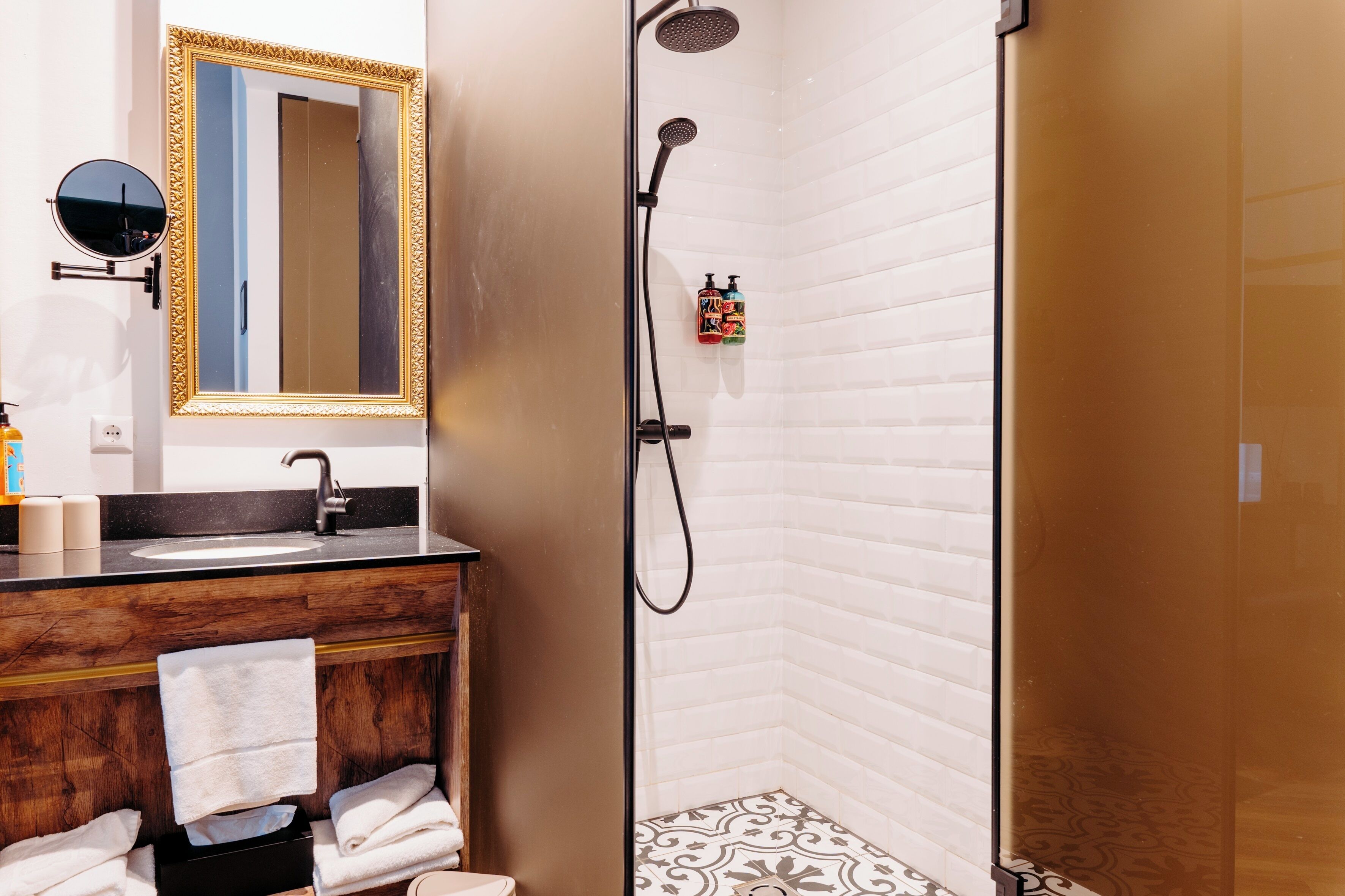 Un baño con un tocador de madera, un espejo redondo con marco dorado, un lavamanos, un perchero y una ducha con paredes de azulejos blancos y suelo estampado.