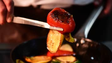 Una persona usa delle pinze per sollevare un pomodoro alla griglia da una padella piena di diversi ortaggi.