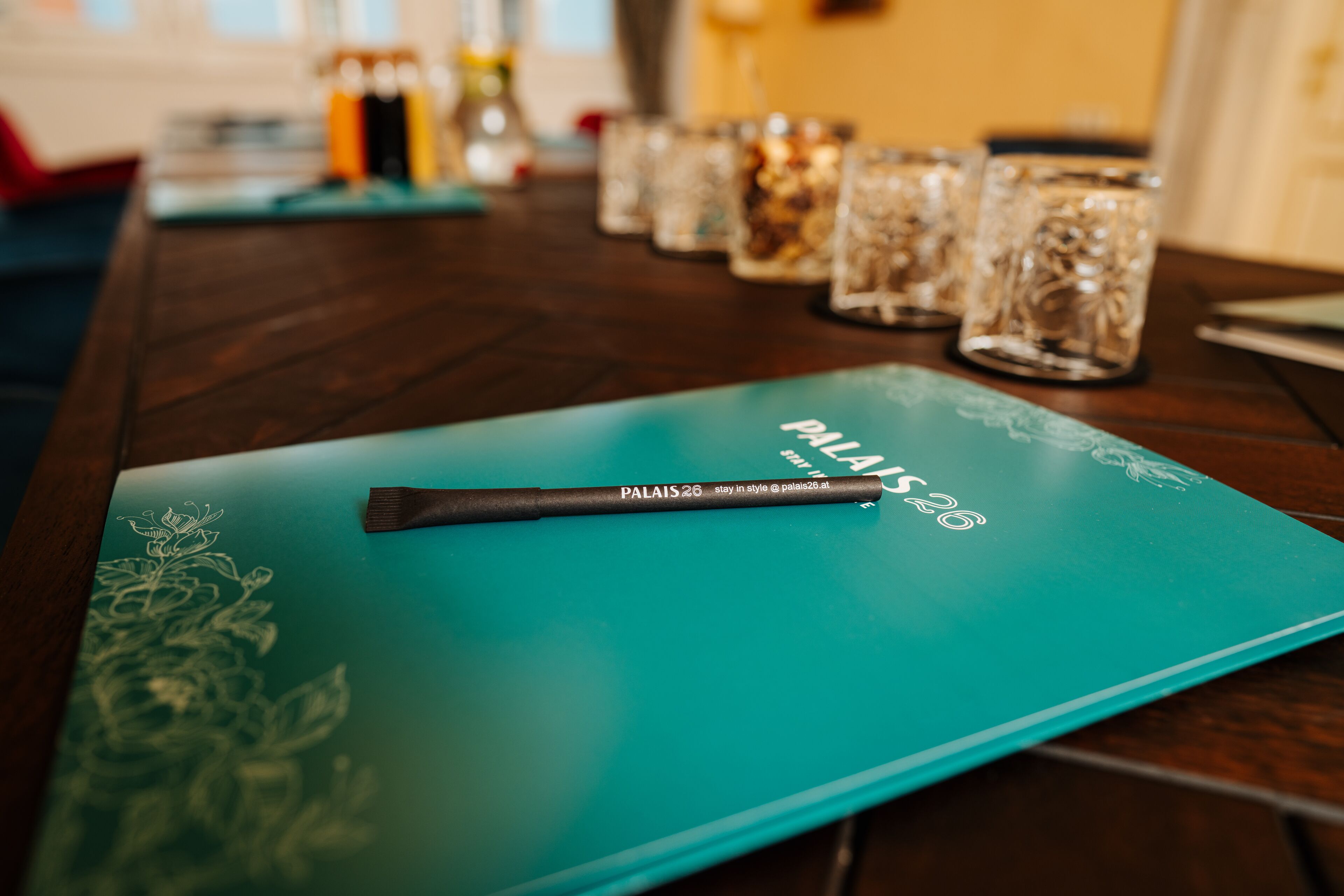A teal-colored tablet on a wooden table with a pen and a glass with a floral design.