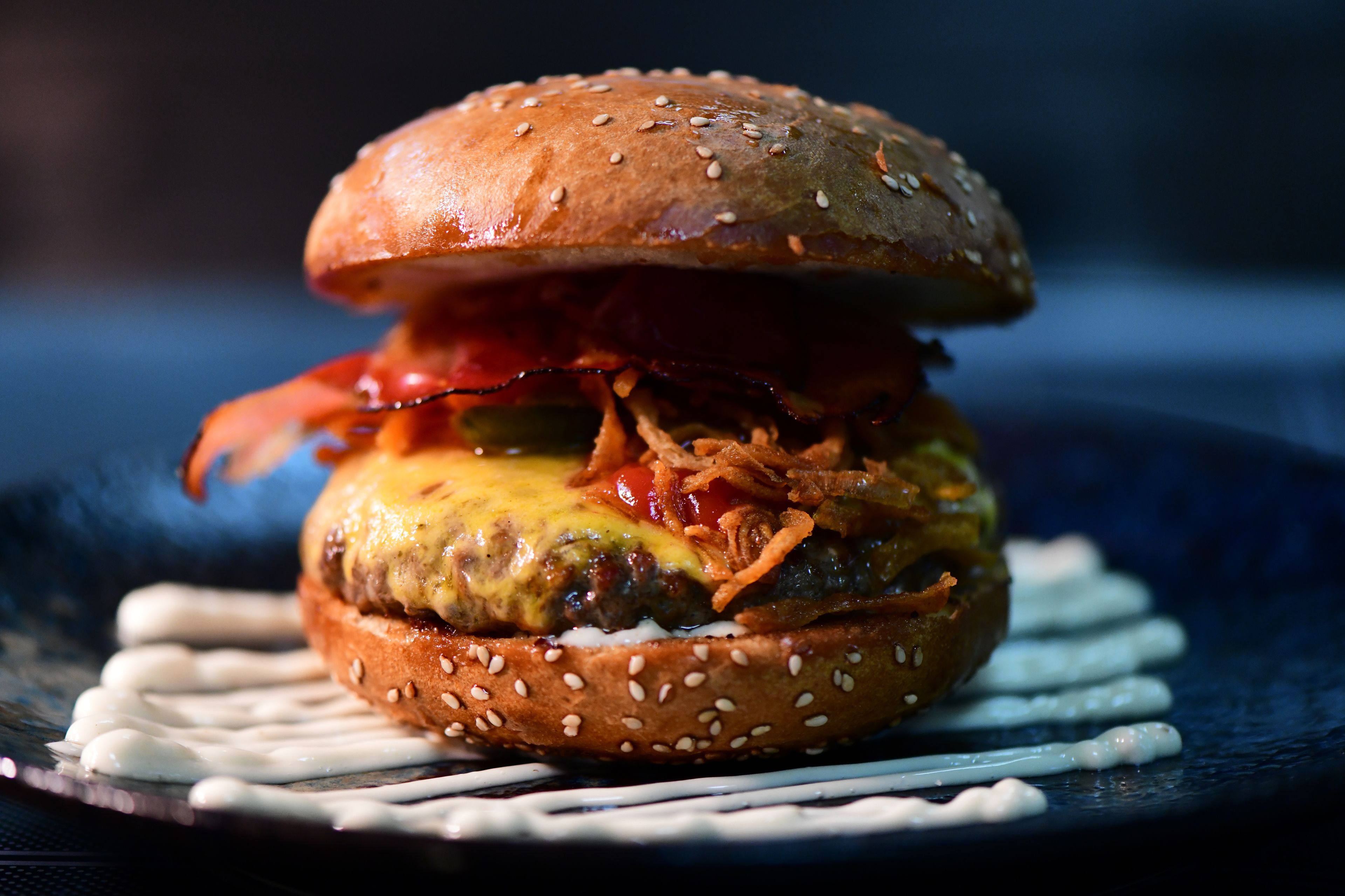 A burger with sesame seed bun, cheese, bacon, and a sauce drizzle.
