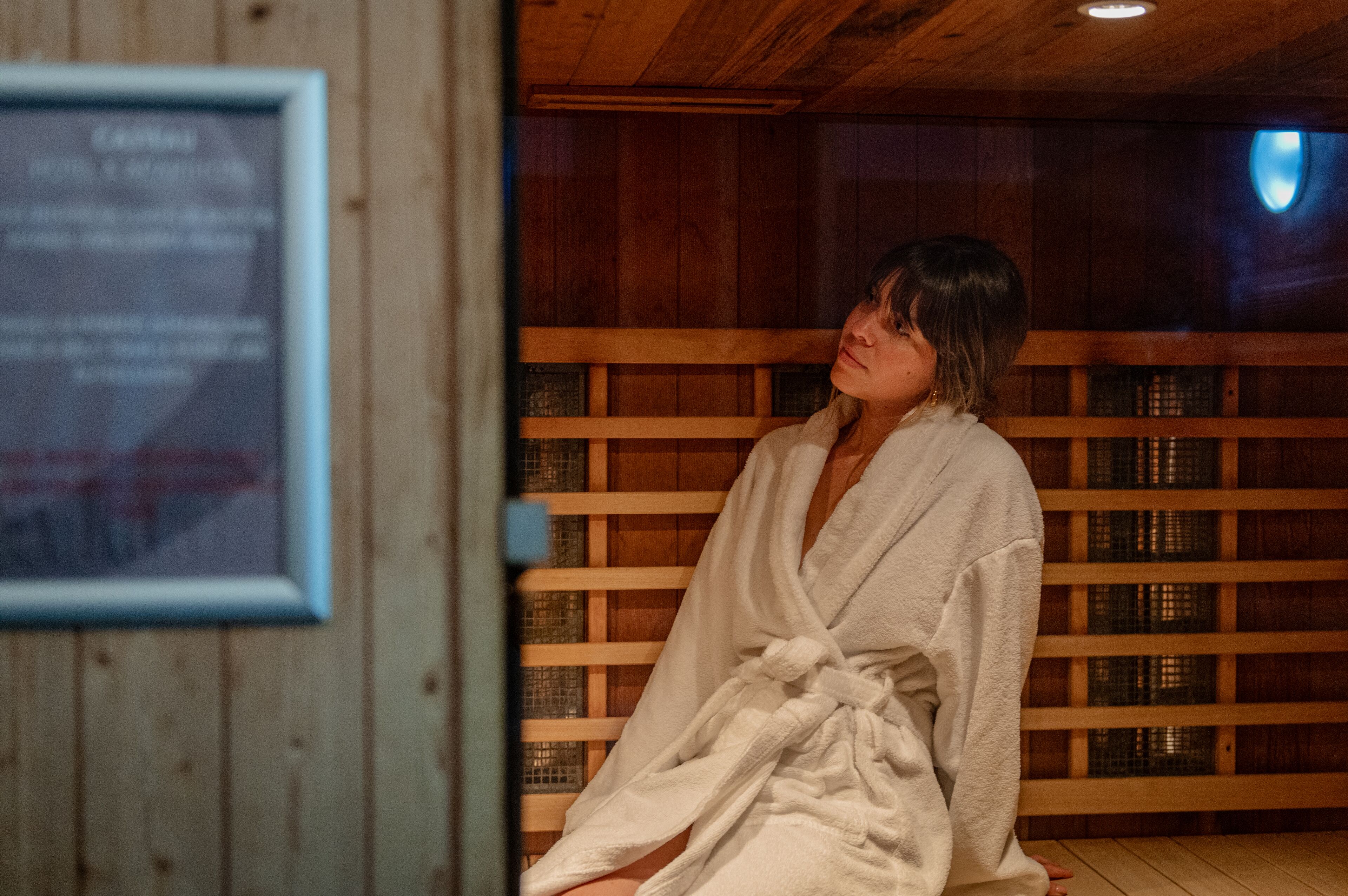 A person in a white bathrobe sitting in a wooden sauna room with a framed sign on the wall.