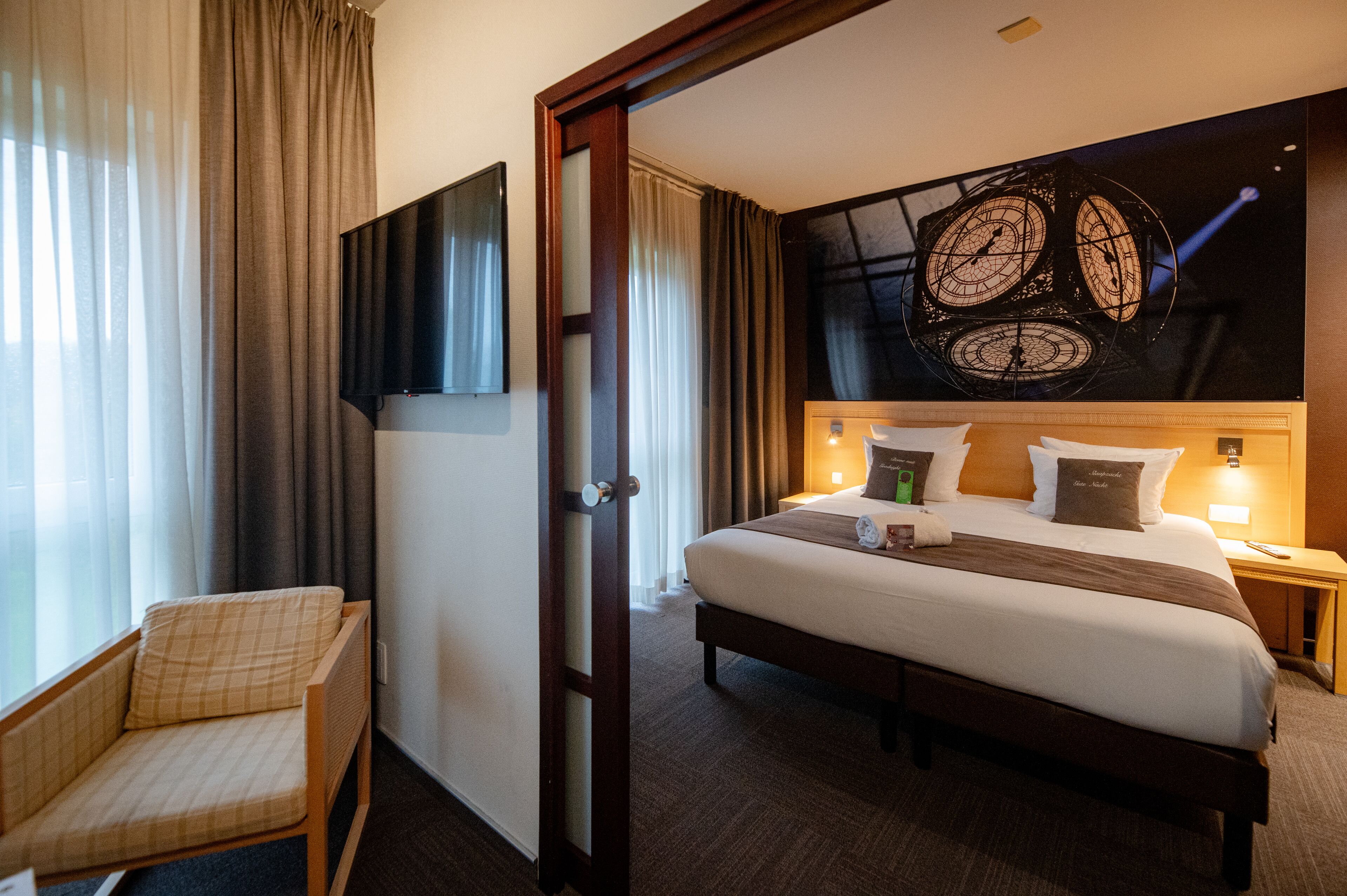 Habitación de hotel con cama, sillón, televisor montado en la pared y un reloj grande.