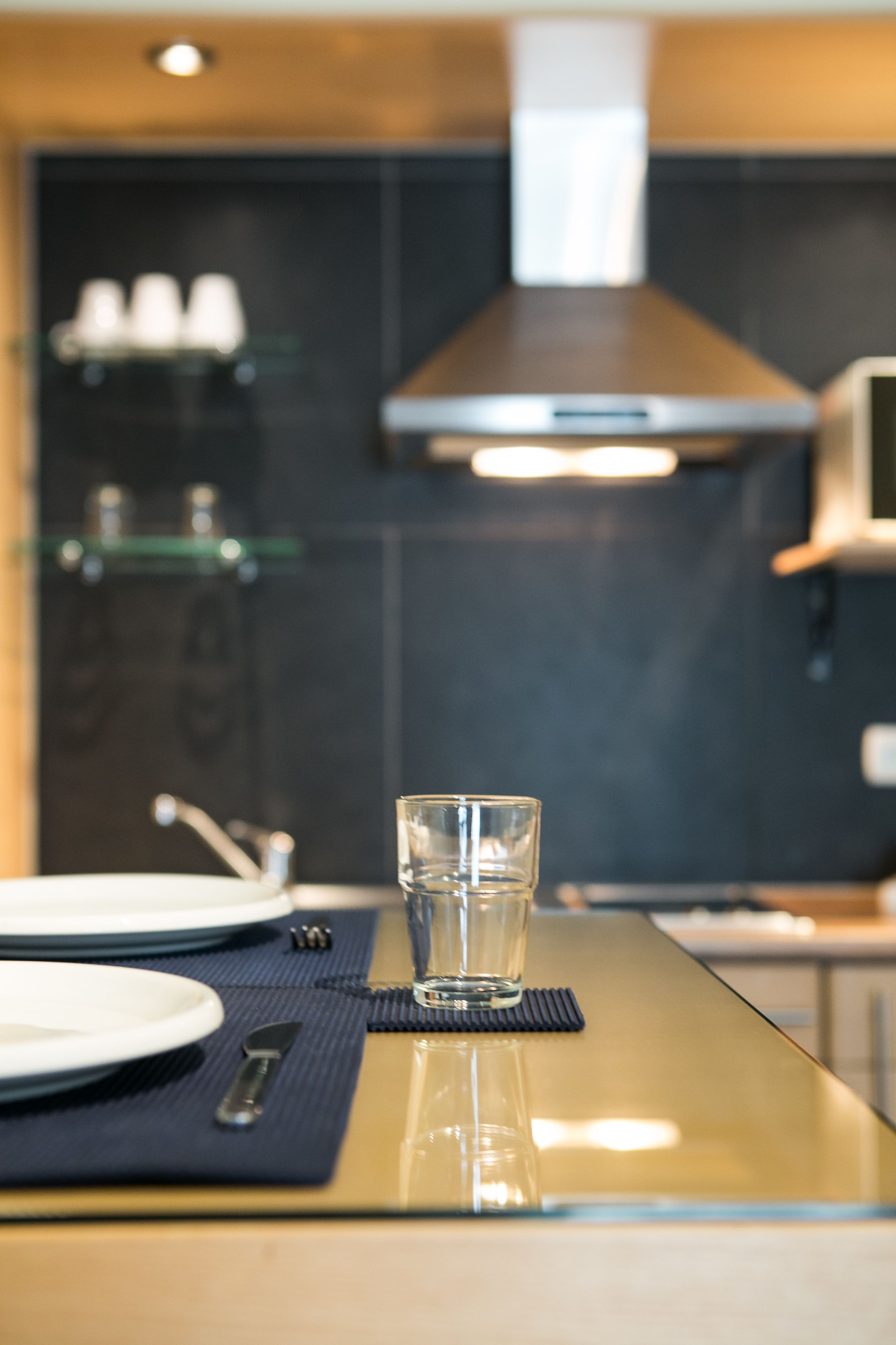 A modern kitchen with a glass-top dining table, a glass of water, and a set of plates and cutlery.