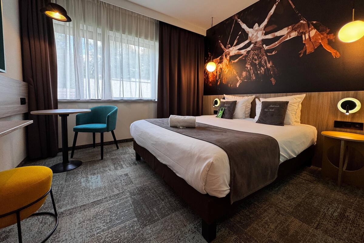 A modern hotel room with a large bed, a desk, a chair, and a large window with curtains.
