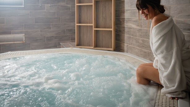 Iemand zit in een bubbelbad, met een betegelde muur en een houten plankje op de achtergrond.