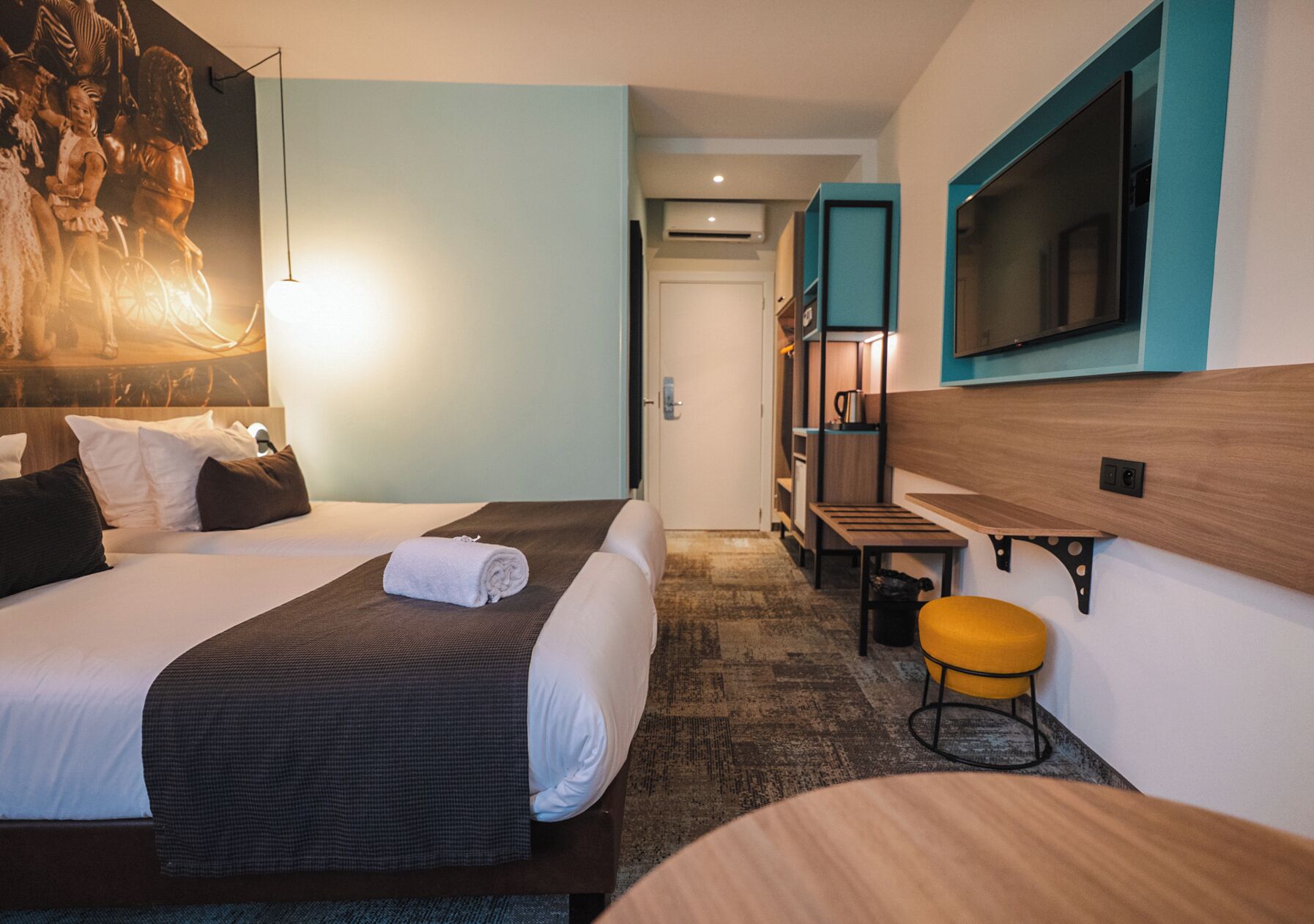 Habitación de hotel con dos camas, un televisor, una mesita pequeña y un taburete amarillo.