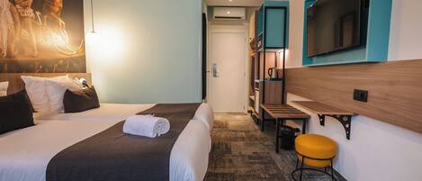 Habitación de hotel con dos camas, un televisor, una mesita pequeña y un taburete amarillo.