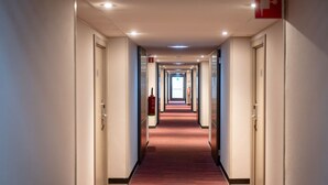 Een hotelgang met een roodgekleurd tapijt, deuren aan weerszijden en zichtbaar brandveiligheidsmateriaal.