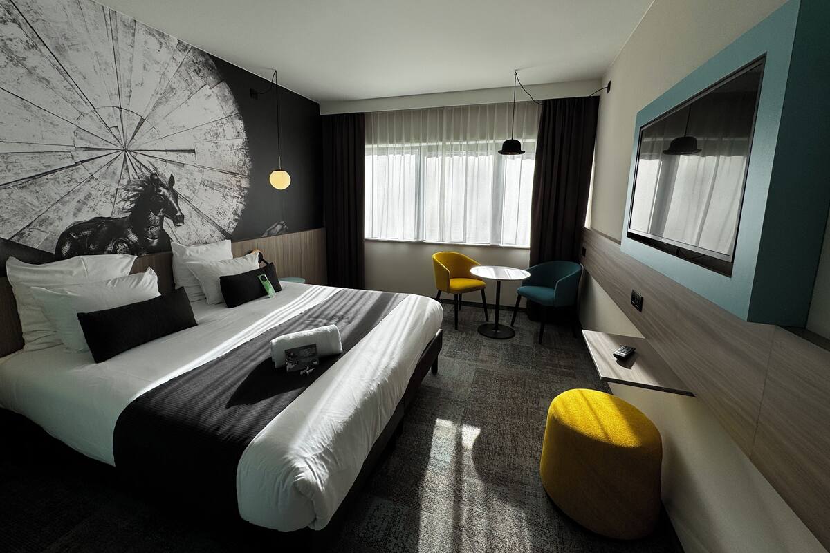 A modern hotel room with a large bed, a small round table, two chairs, a yellow ottoman, and a large window with curtains.
