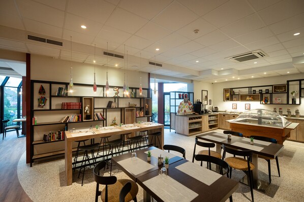 A modern cafe with a long communal table, individual tables, and a variety of seating options. - Hotel & Aparthotel Casteau Resort Mons (Soignies)