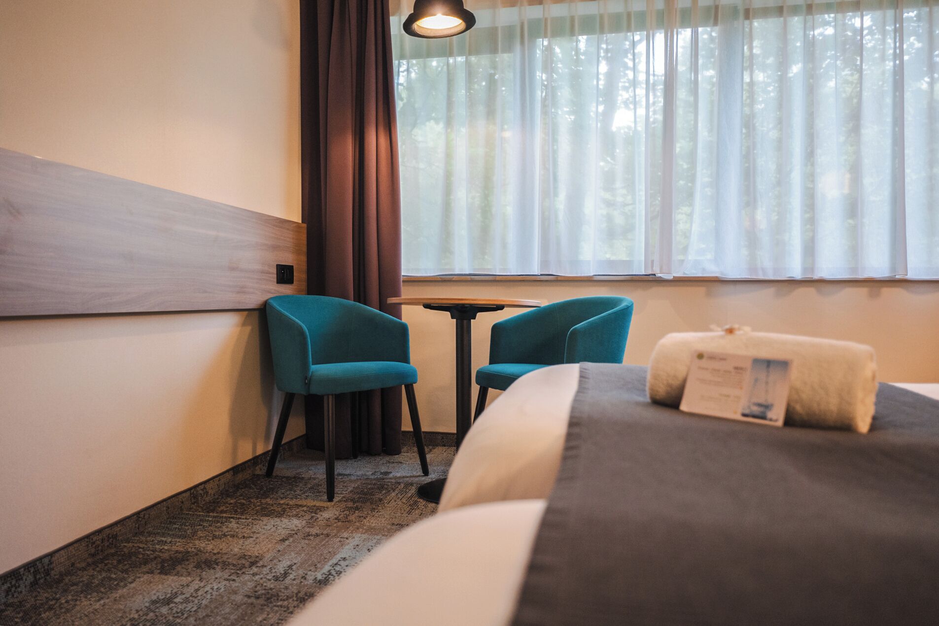 A hotel room with a bed, two blue chairs, a small table, and a window with sheer curtains.