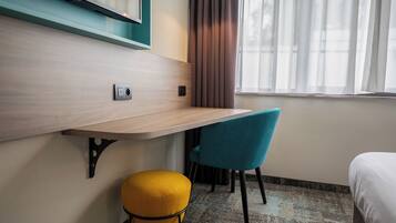 A hotel room with a wooden desk, a blue chair, a yellow ottoman, a bed, a window with curtains, and a wall-mounted light fixture.