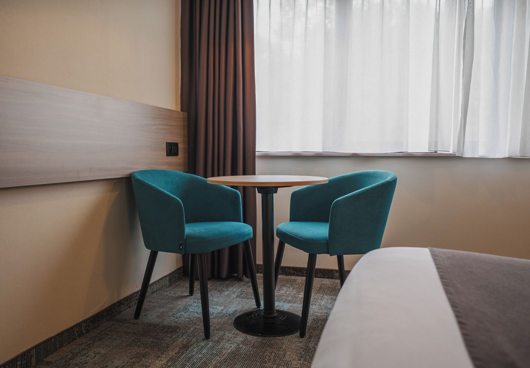 Une chambre d’hôtel avec un lit, deux fauteuils turquoise, une petite table ronde, une fenêtre avec des rideaux et un panneau fixé au mur.