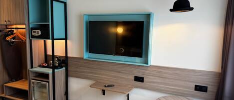 A modern hotel room with a large flat-screen TV mounted on the wall, a small wooden table, a yellow stool, a black trash bin, and a coat rack with a hanging garment.