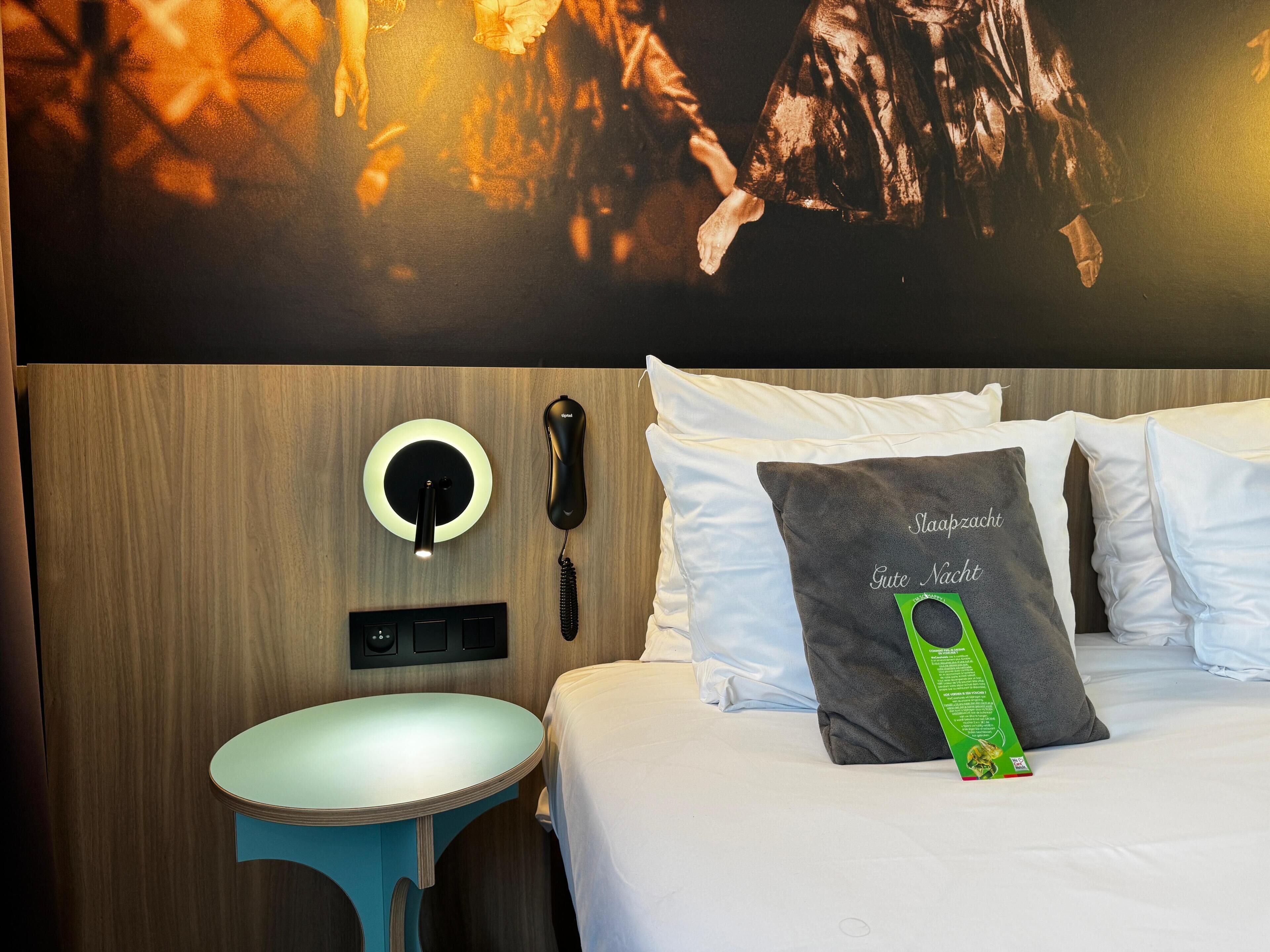 A hotel room with a bed, a bedside table, a wall-mounted phone, and a wall art piece.