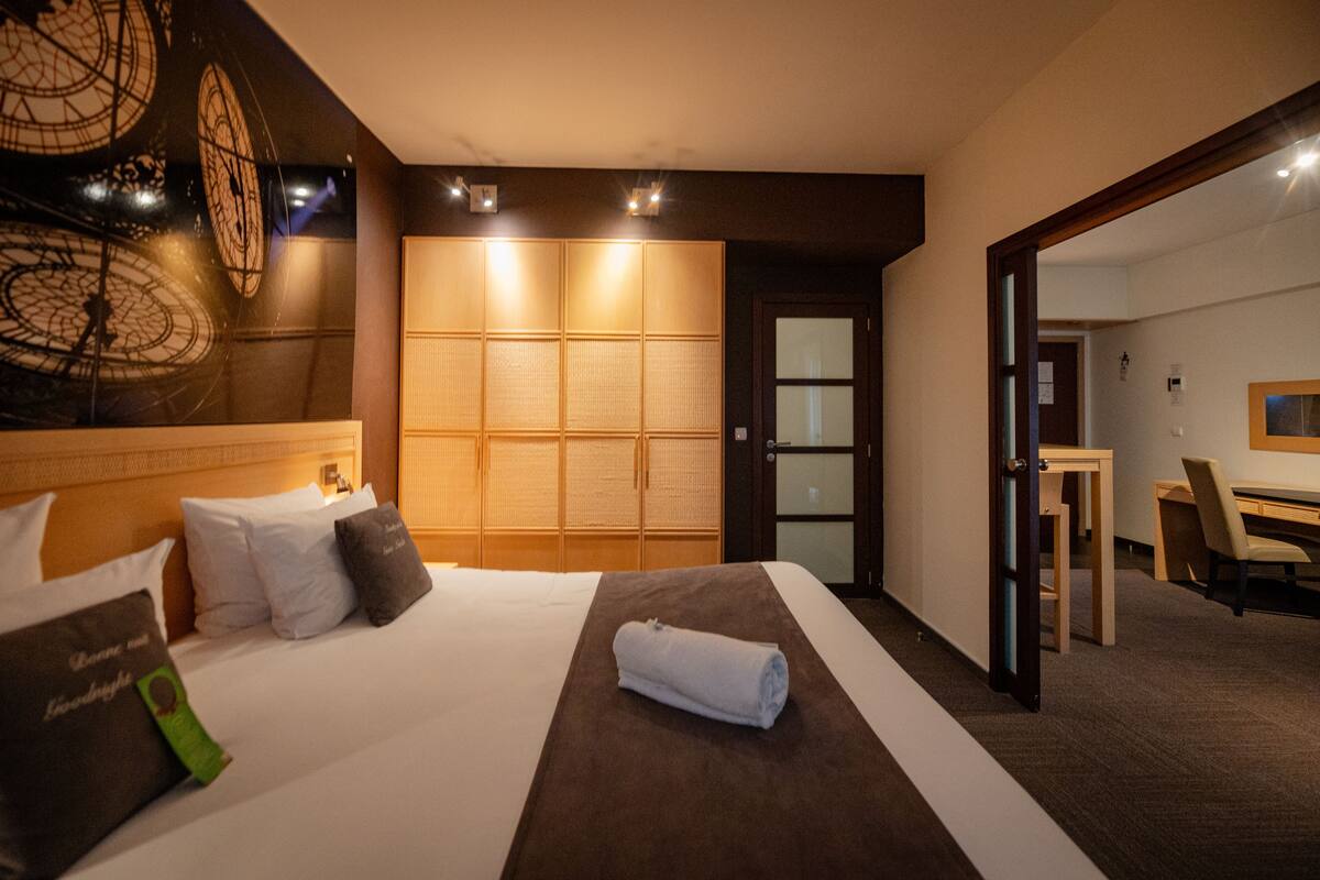 A hotel room with a large bed, a wooden wardrobe, a desk with a chair, and a mirror.