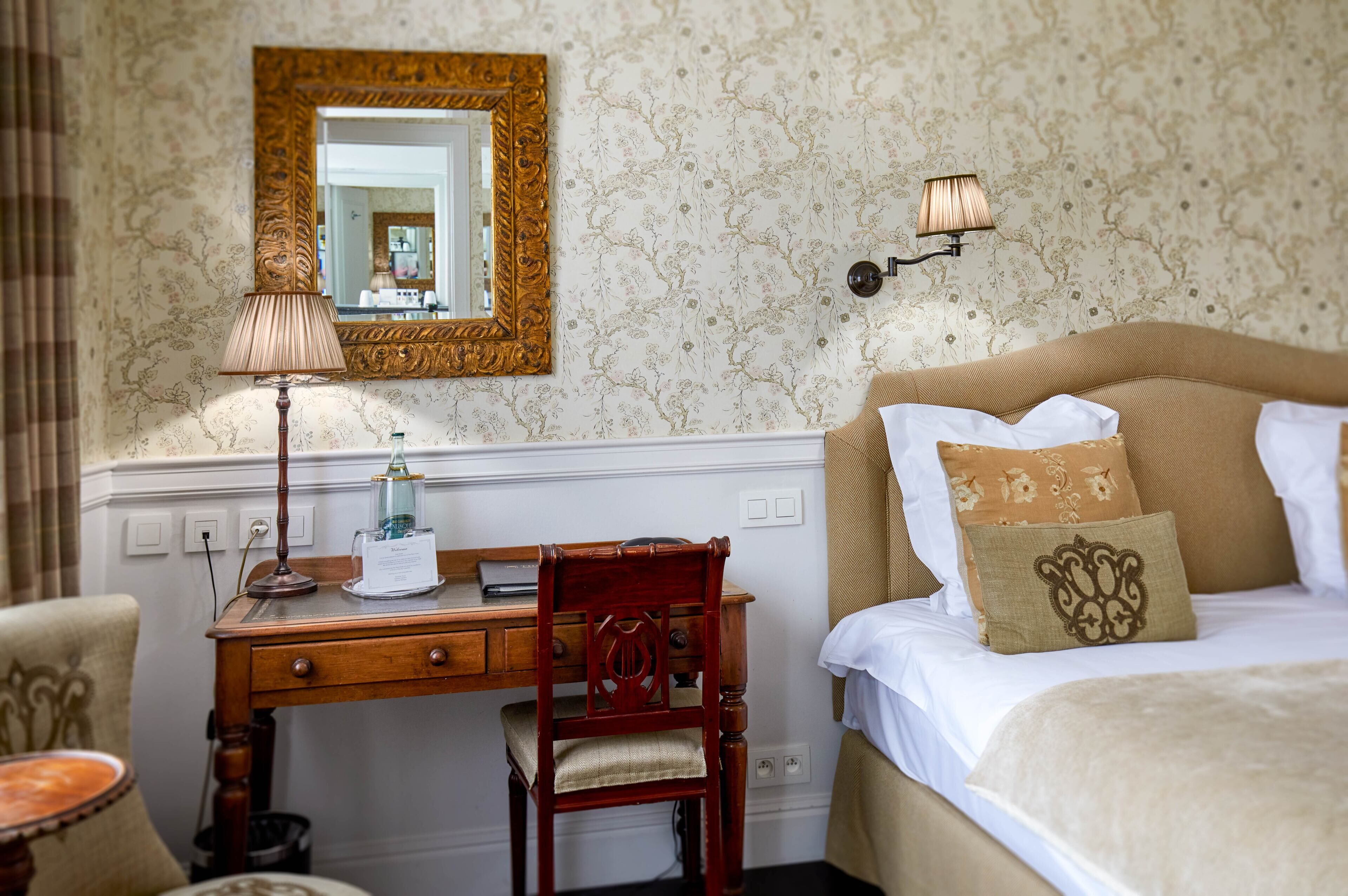 A bedroom with a bed, a desk, a chair, a lamp, a mirror, and a wall with floral wallpaper.