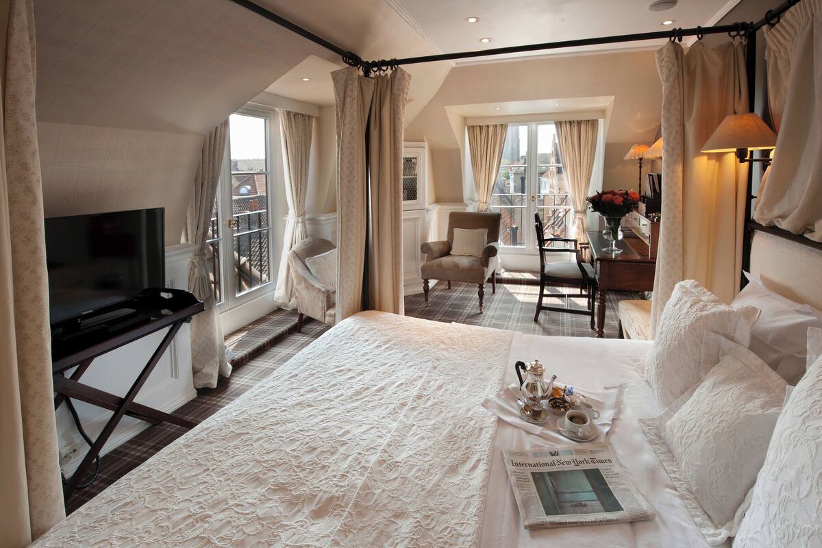 A hotel room with a large bed, a television, a dining area with a table and chairs, and a view of the city through the windows.