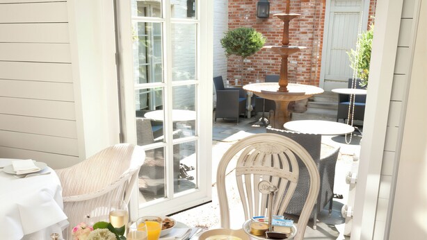 Una mesa de desayuno dispuesta con diversos platos, entre ellos frutas, pasteles y bebidas, en un patio luminoso y abierto con vistas a un edificio de ladrillo y mobiliario exterior.
