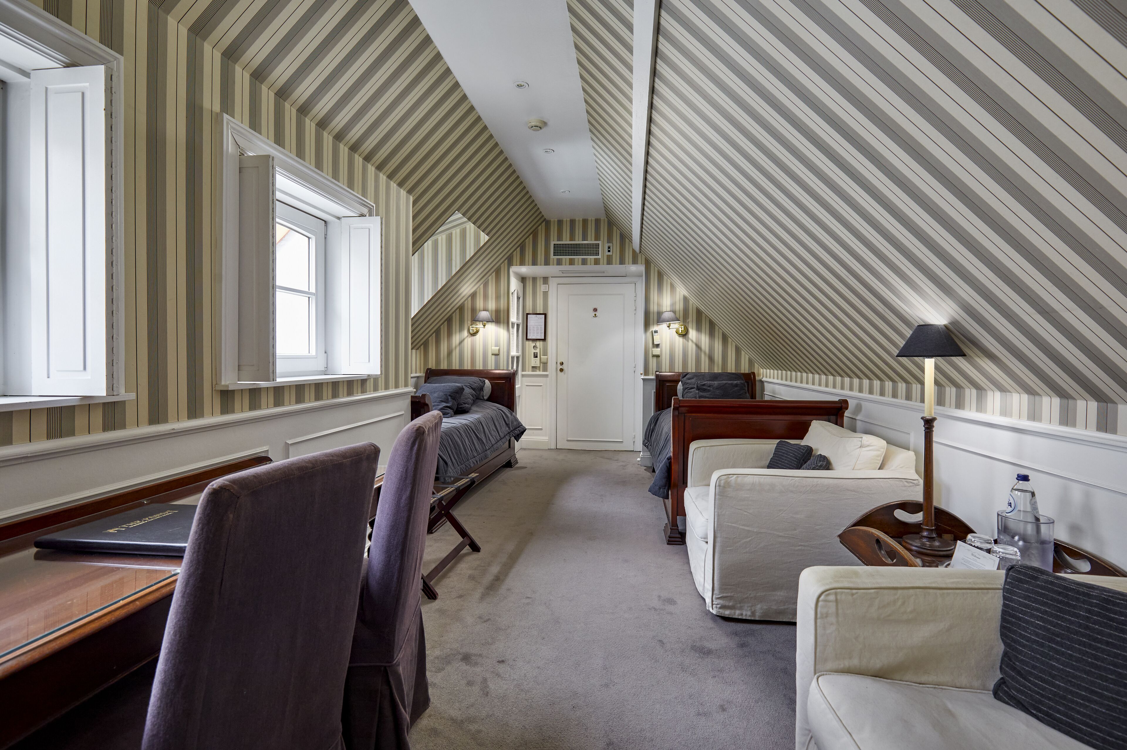 Hotelkamer met een schuine plafond, gestreepte behang, een bureau met een laptop, twee bedden, een stoel, een lamp en een waterfles.