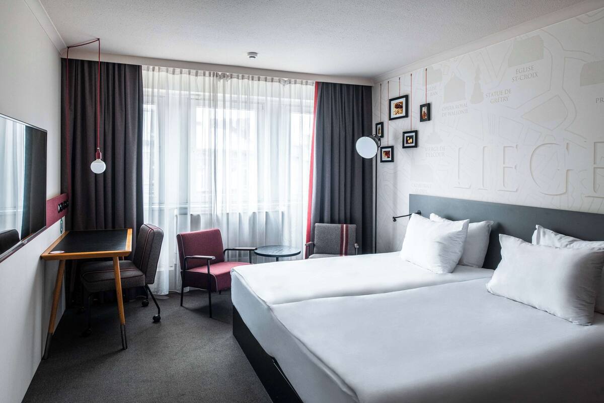 Quarto de hotel com duas camas, uma escrivaninha, uma cadeira, uma mesinha e uma janela com cortinas.