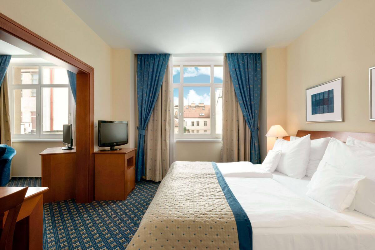 Une chambre d’hôtel avec un lit, un téléviseur posé sur un meuble en bois, un bureau et une fenêtre avec des rideaux.