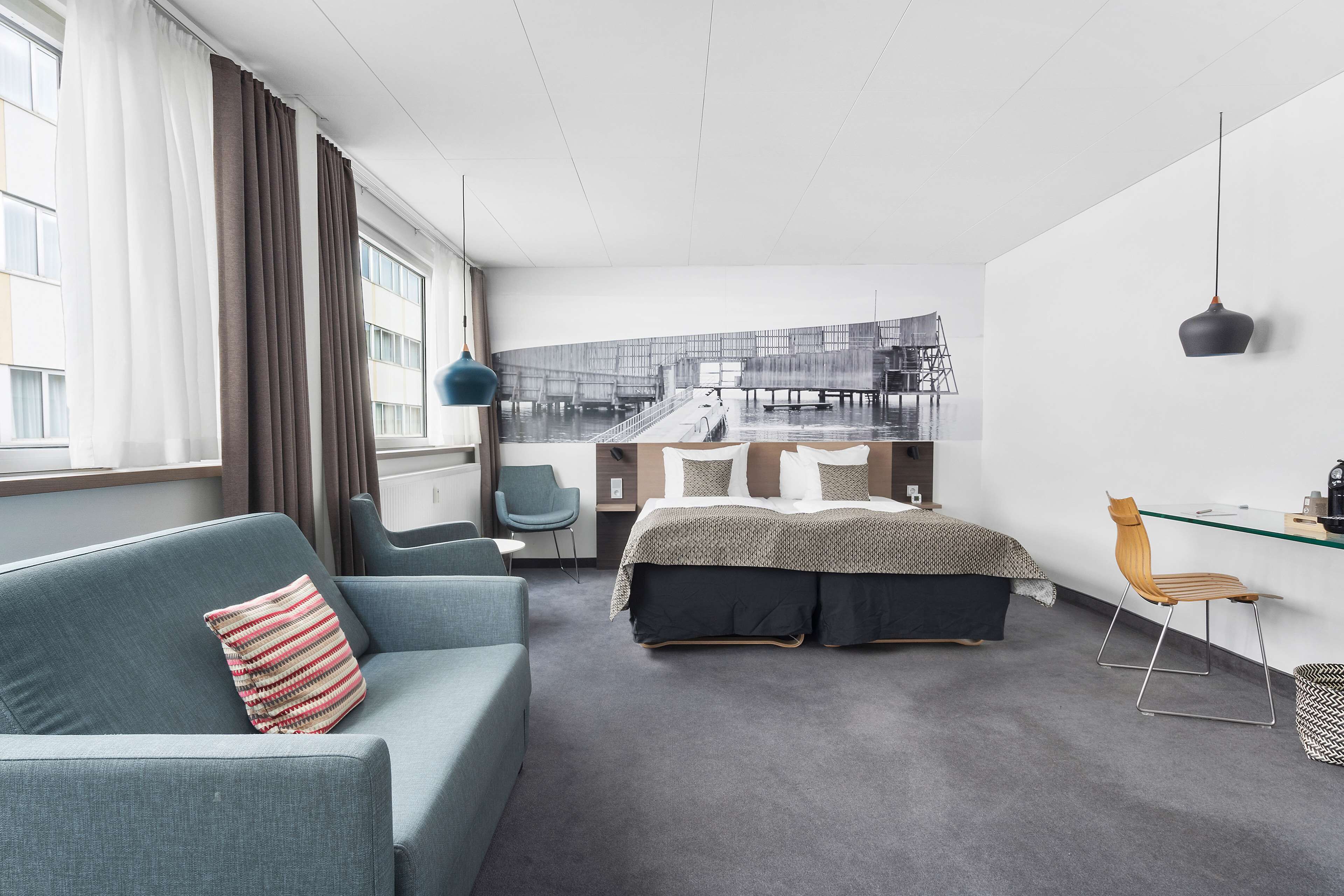A modern hotel room with a grey sofa, a bed with a patterned bedspread, a desk with a chair, and a large window with curtains.