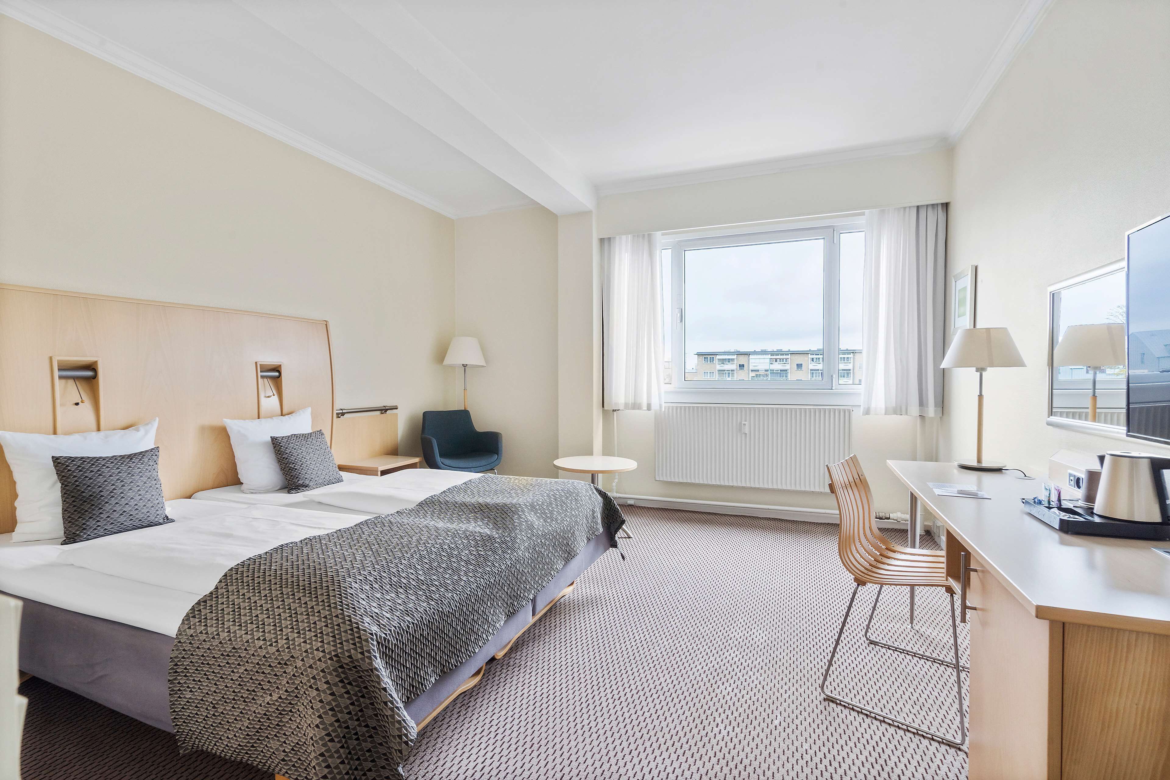Une chambre d’hôtel avec un grand lit, un bureau avec une chaise, une fenêtre avec des rideaux et une vue sur les bâtiments environnants.