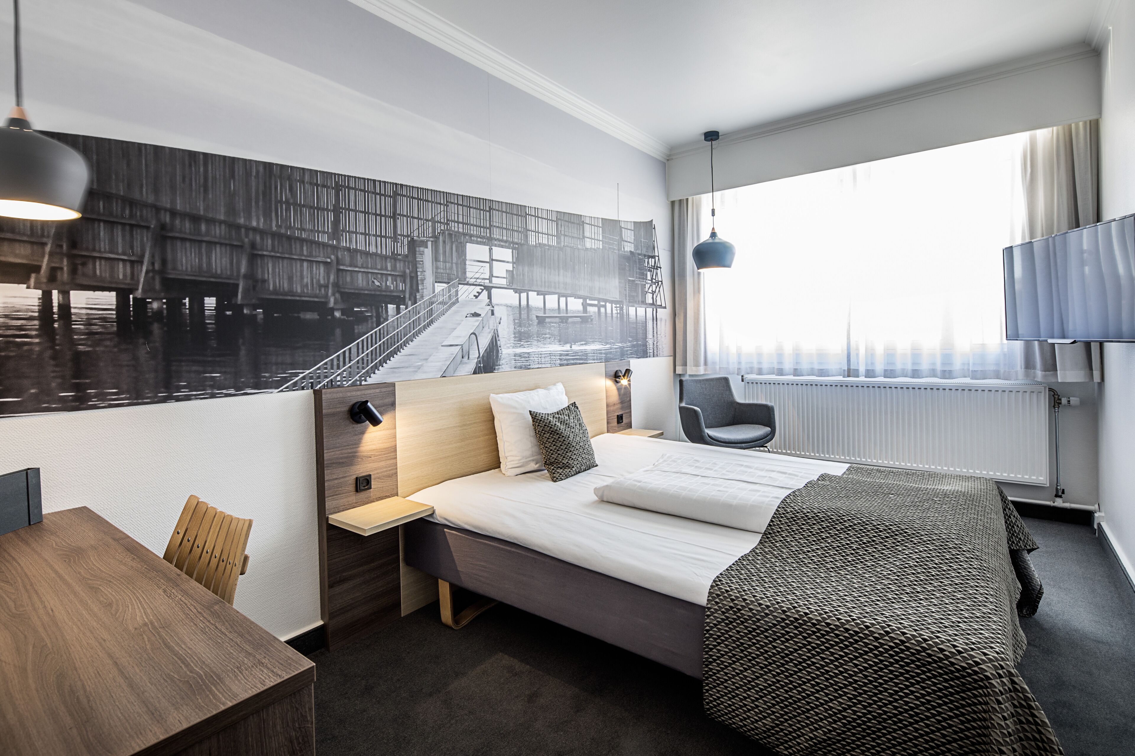 A modern hotel room with a large bed, a desk, and a chair. There is a large window with a view of a cityscape.