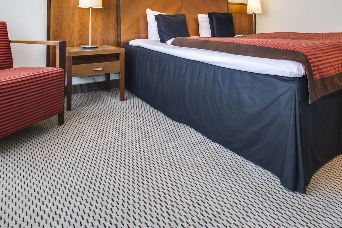Une chambre d’hôtel avec un lit, deux tables de chevet avec des lampes, une tête de lit en bois, une chaise rouge et un tapis à motifs.