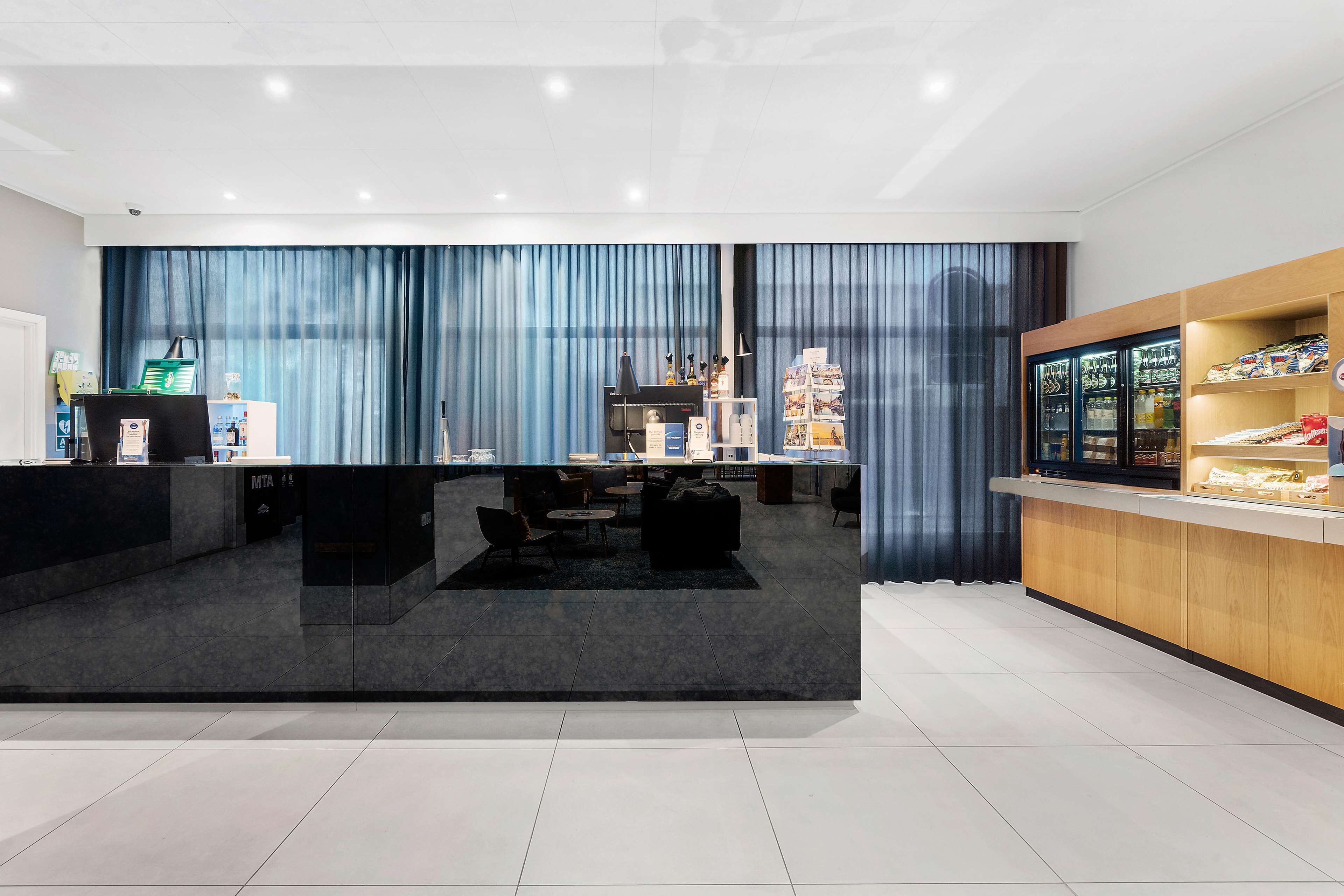 A modern hotel lobby with a reception desk, seating area, and a refreshment bar.