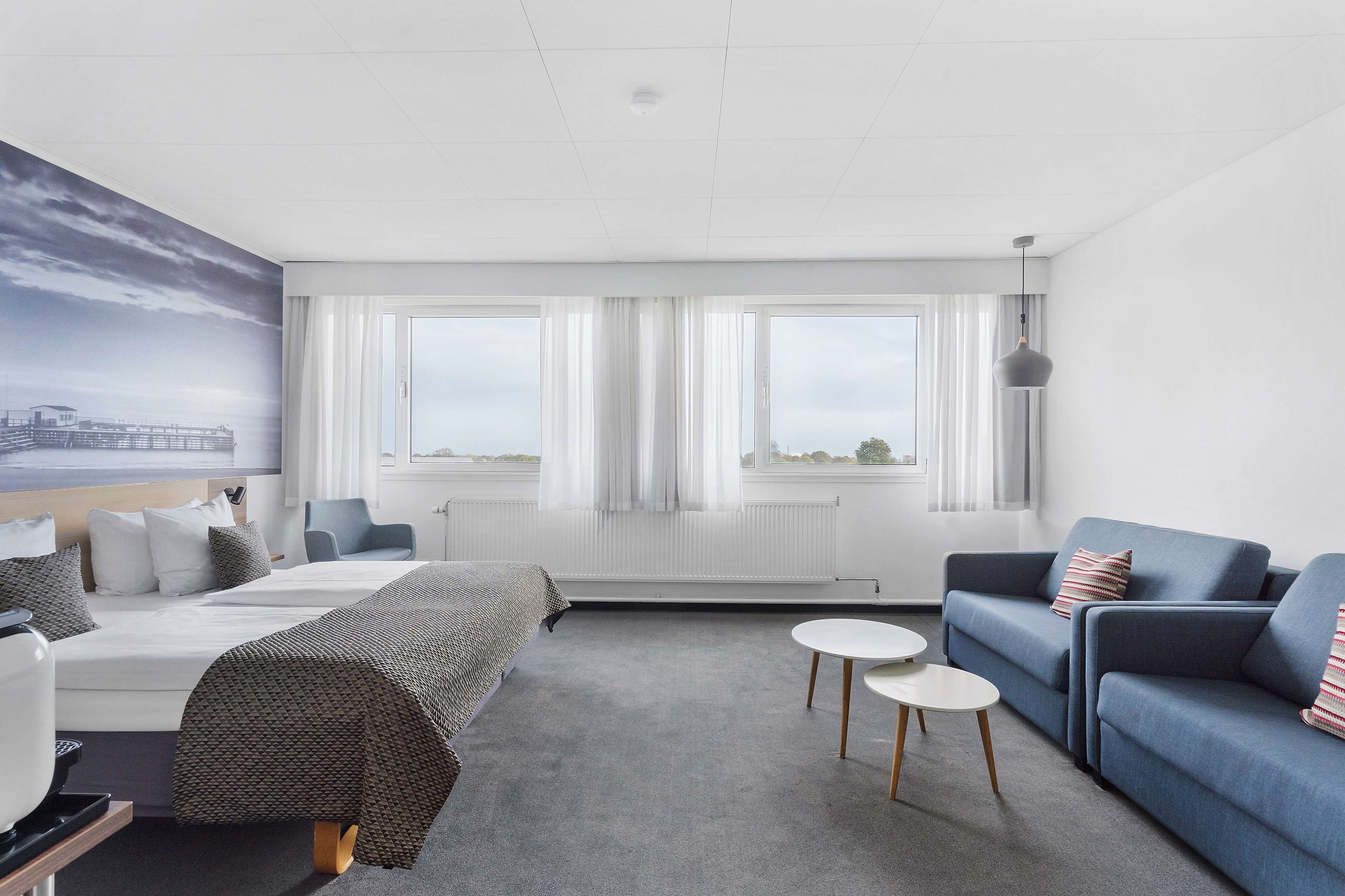 A modern hotel room with a bed, a blue sofa, a small white table, and large windows with curtains.