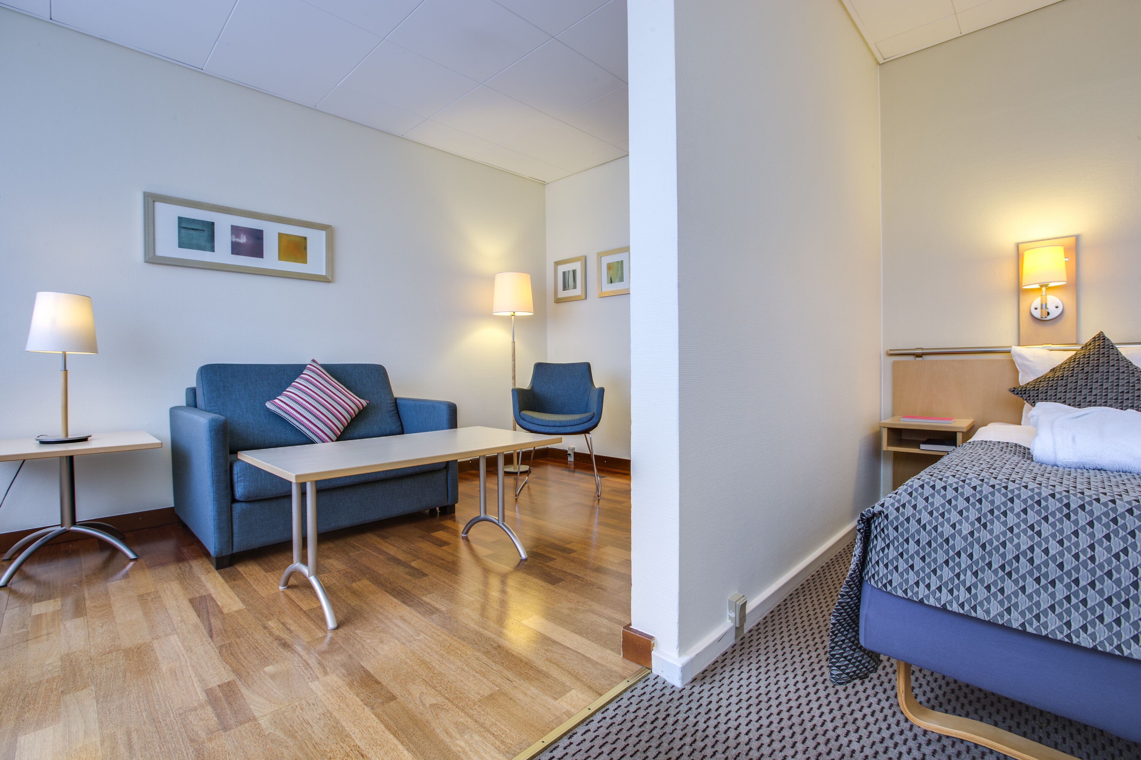 A hotel room with a bed, a sofa, a chair, a small table, and two lamps.