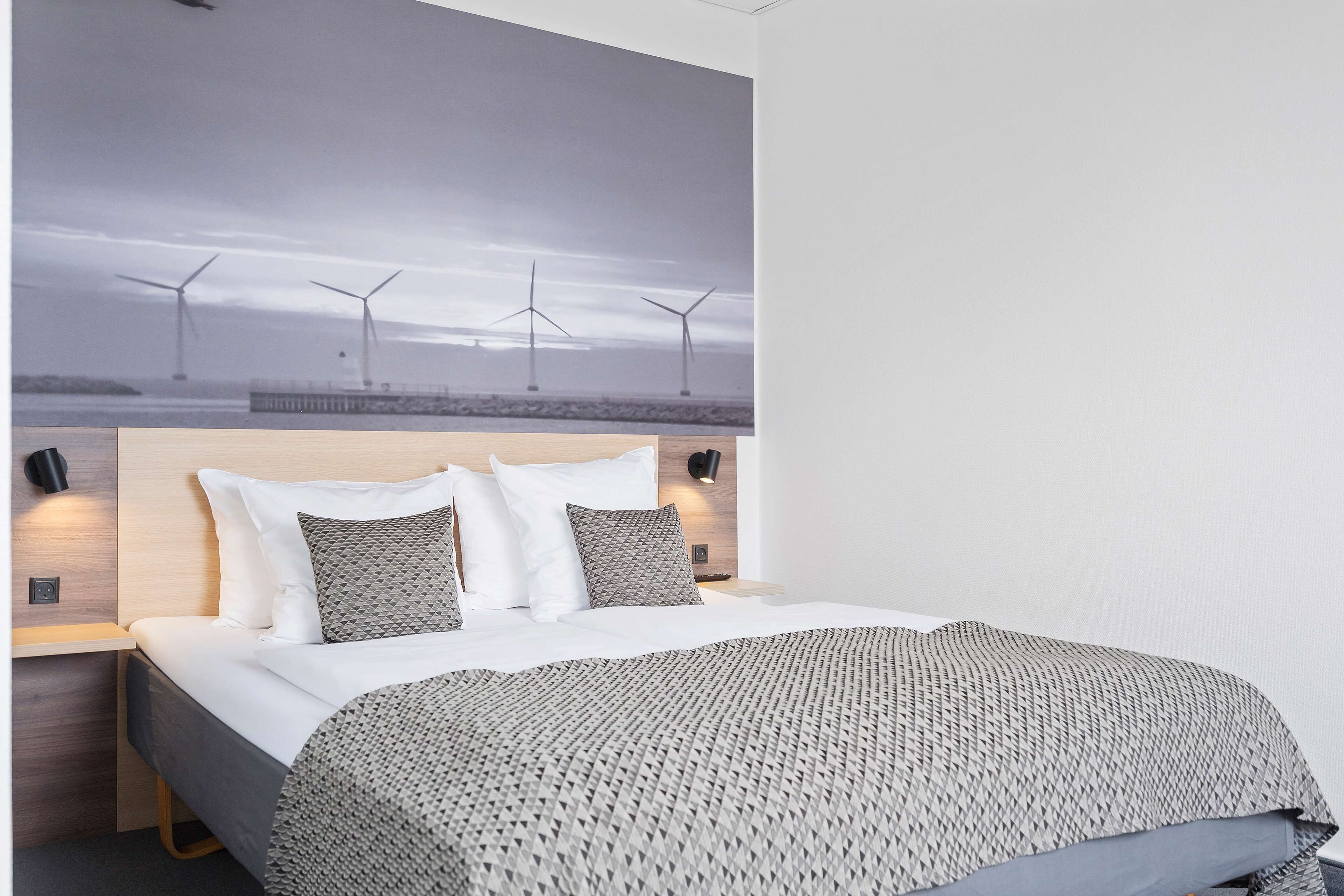 A hotel room with a bed, a headboard with a large picture of wind turbines, and bedside lamps.